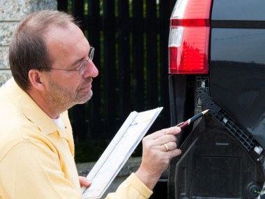 Kfz-Gutachter zeigt mit einem Stift auf den Schaden am Heck des Autos und hebt ein Klemmbrett für Notizen